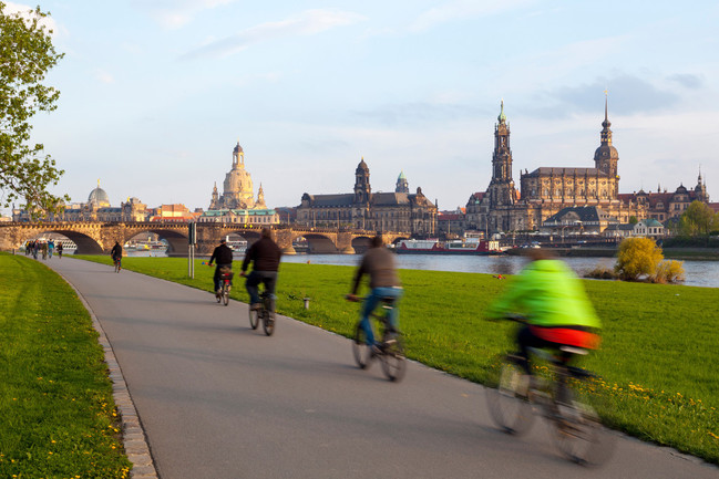 Radfahrer fahren auf dem Radweg am Dresdner Elbufer unterhalb des Japanischen Palais vor der abendlichen Kulisse der Dresdner Altstadt.